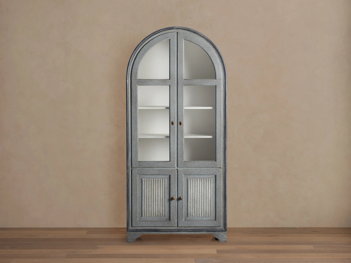 Giotto Cabinet in antique blue with arched top, glass doors with brass handles, and visible interior shelving, against beige wall.