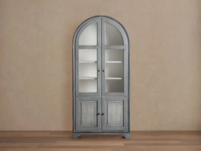 Giotto Cabinet in antique blue with arched top, glass doors with brass handles, and visible interior shelving, against beige wall.