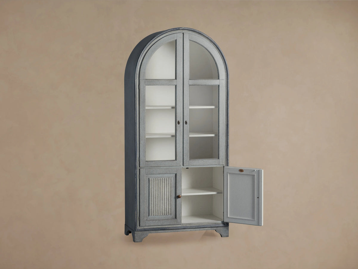 Giotto Cabinet in antique blue with arched top, glass doors with brass handles, and visible interior shelving, against beige wall.
