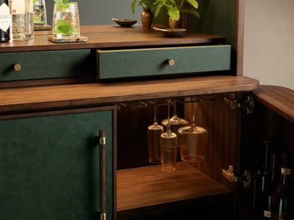 Close-up of the forest green and walnut Grant Bar Cabinet, an open drawer, brass knobs, hanging stemware, and stored wine bottles.