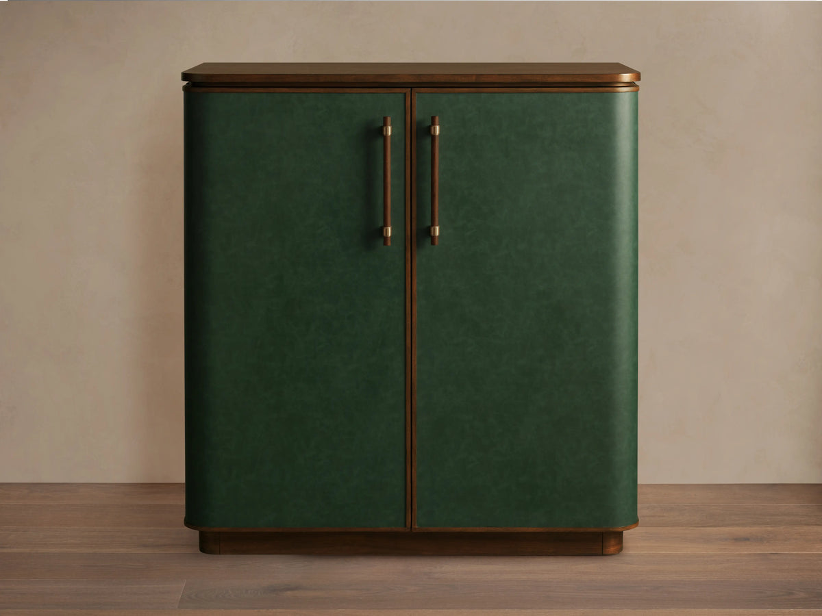 The Grant Bar, crafted from walnut wood and green faux leather with brass handles, stands on a wooden floor against a neutral wall.