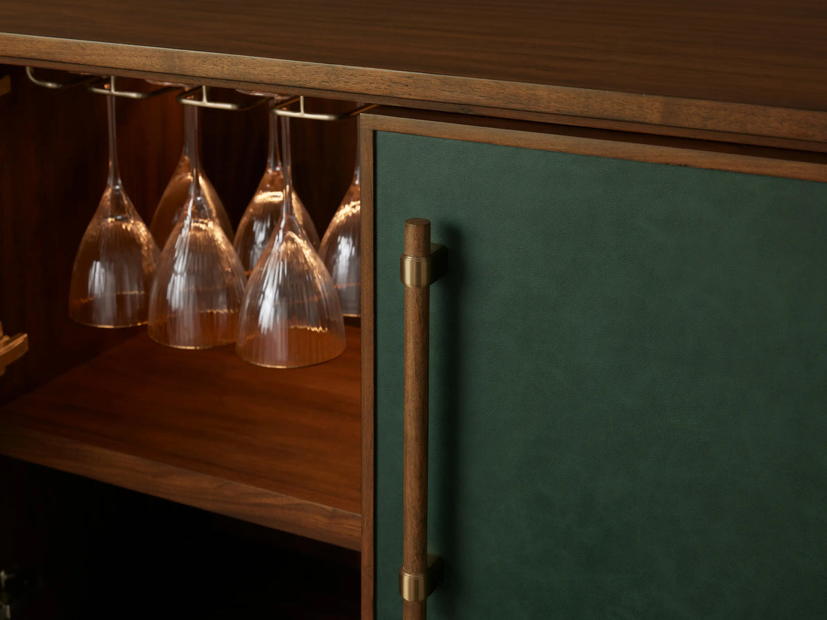 The Grant Bar, crafted from walnut wood and green faux leather with brass handles, stands on a wooden floor against a neutral wall.