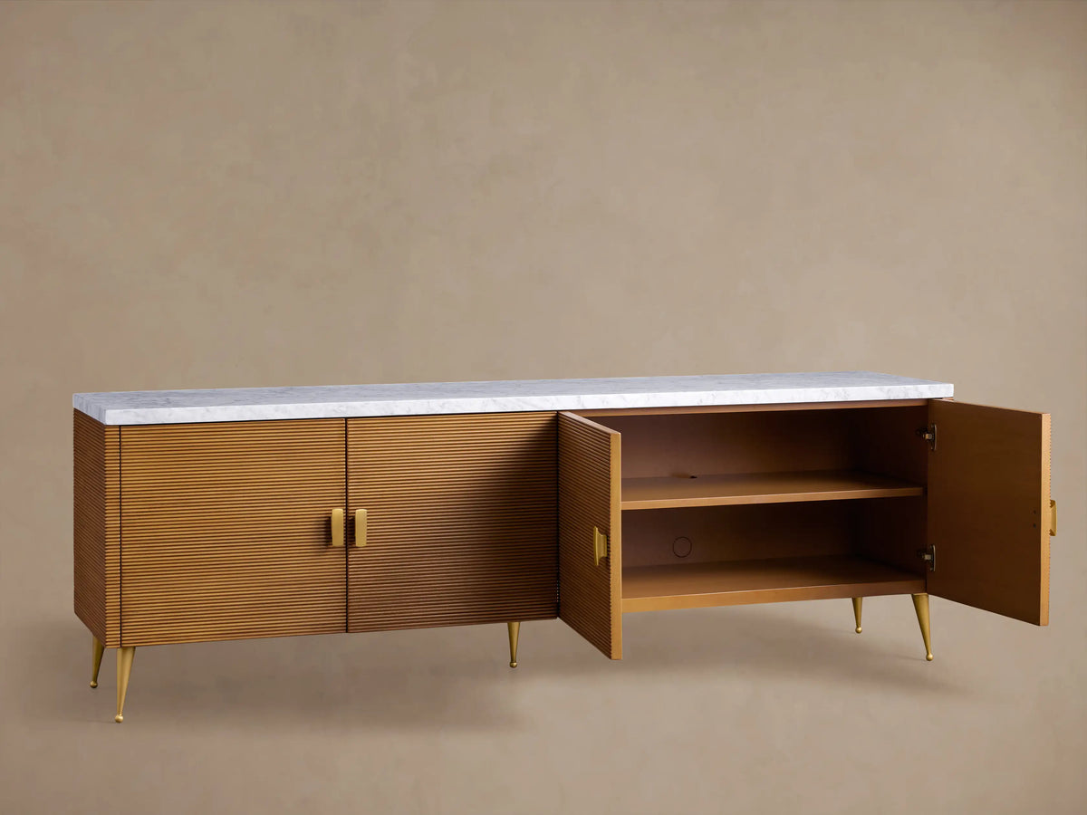 Giselle Media Console with reeded warm brown doors, white marble top, and brass handles and legs, against a tan wall.