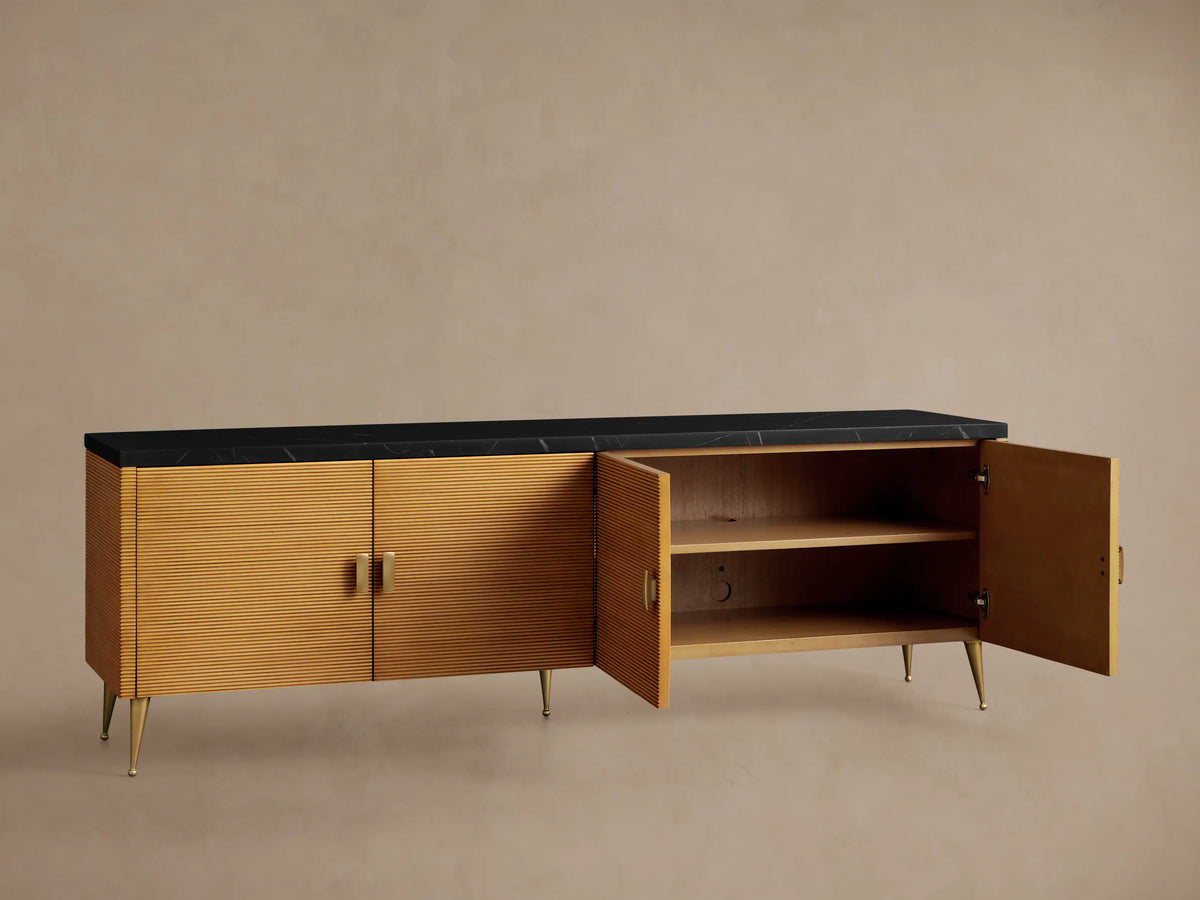 Giselle Media Console: long, horizontal, golden-brown wood body, 4 ribbed doors, black marble top, brass handles & tapered legs.