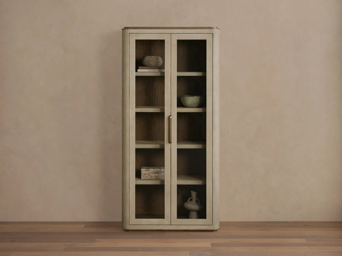 Harrow Cabinet: Tall, light beige faux shagreen cabinet with rounded edges, two glass doors, brass handle, and wooden shelves displaying decorative objects.