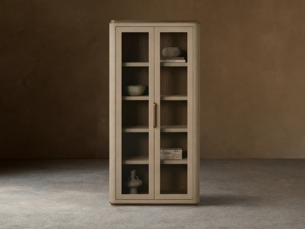 Harrow Cabinet: Tall, light beige faux shagreen cabinet with rounded edges, two glass doors, brass handle, and wooden shelves displaying decorative objects.