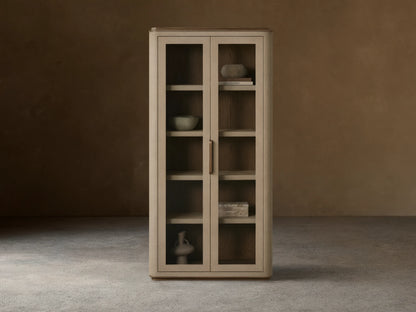 Harrow Cabinet: Tall, light beige faux shagreen cabinet with rounded edges, two glass doors, brass handle, and wooden shelves displaying decorative objects.