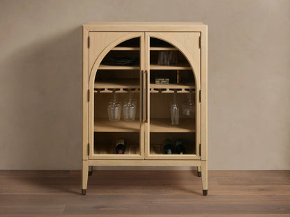 Hattie Bar Cabinet in light wood with glass doors, reeded hardware, and brass-capped feet. Visible shelves with stemware and bottles.