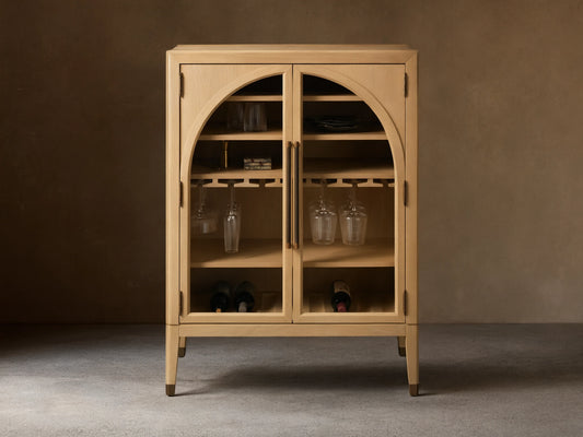 Hattie Bar Cabinet in light wood with glass doors, reeded hardware, and brass-capped feet. Visible shelves with stemware and bottles.