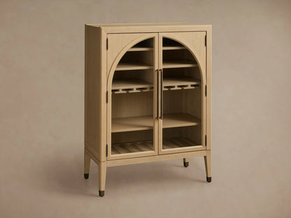 Hattie Bar Cabinet in light wood with glass doors, reeded hardware, and brass-capped feet. Visible shelves with stemware and bottles.