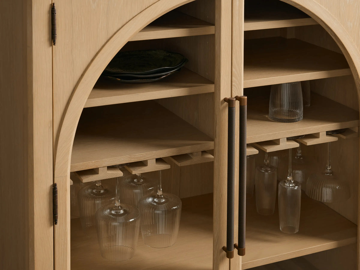 Hattie Bar Cabinet made of oak wood with glass doors, reeded hardware, adjustable shelves, and brass-capped feet on a neutral backdrop.