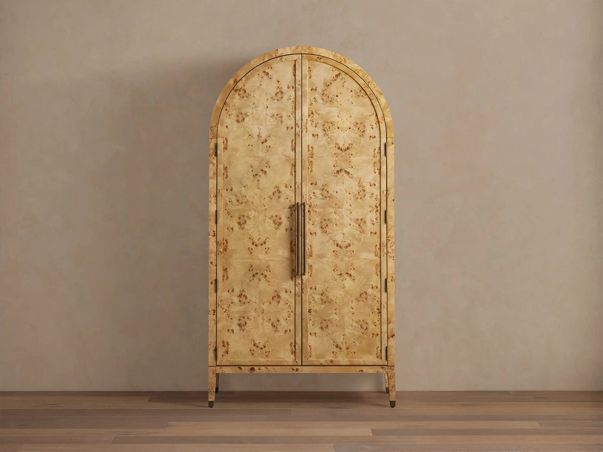 Hattie Burl Cabinet with arched top, swirling mappa burl pattern, brass-capped feet, and vertical reeded handles on closed doors.
