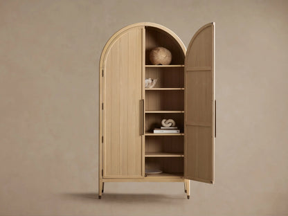Oak wood Hattie Cabinet with an arched top, reeded hardware, and brass-capped feet against a beige wall and wooden floor.