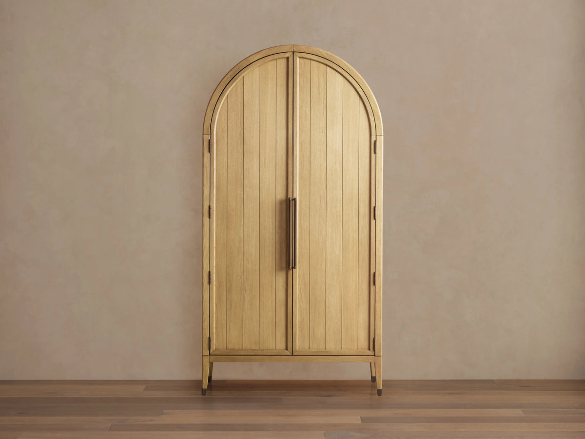 Arched wooden cabinet in light oak finish with vertical panels and antique-style hardware. Set against a plain wall on a wooden floor.