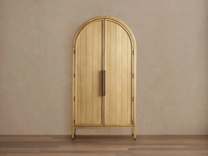 Arched wooden cabinet in light oak finish with vertical panels and antique-style hardware. Set against a plain wall on a wooden floor.
