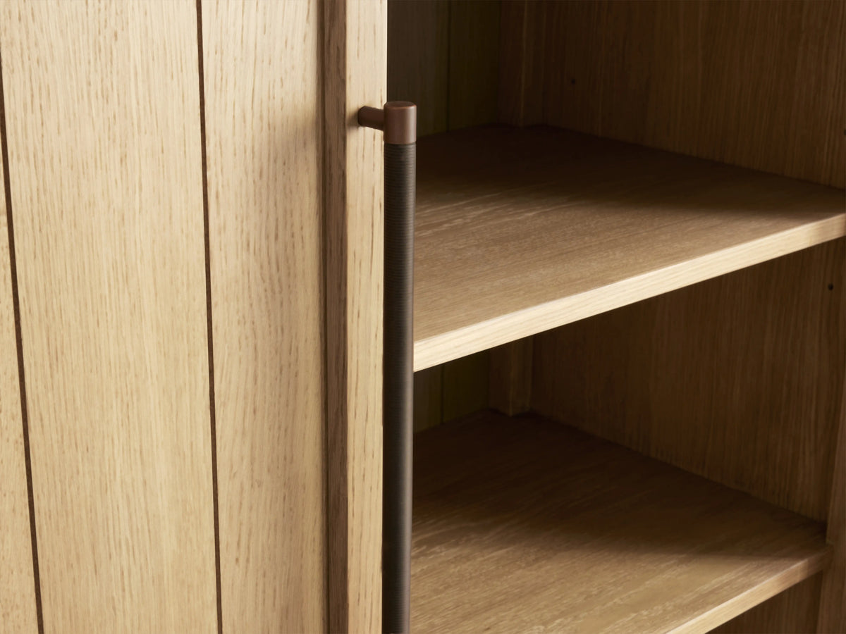 Arched wooden cabinet in light oak finish with vertical panels and antique-style hardware. Set against a plain wall on a wooden floor.