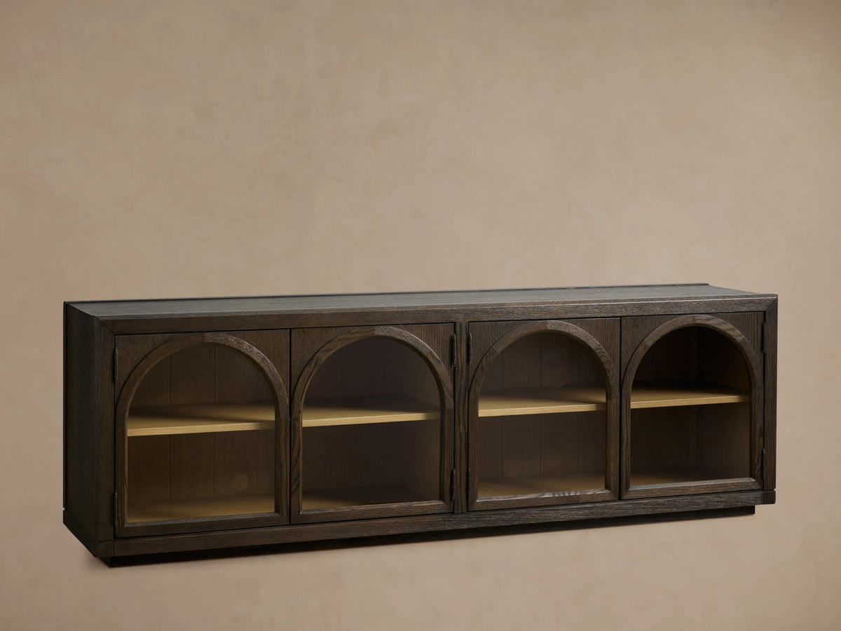 Hattie Media Console made of dark oak wood with three arched doors, revealing shelves, placed against a neutral wall.