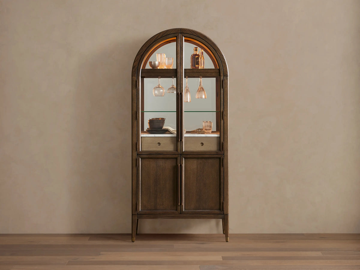 Hattie Bar Cabinet, dark wood with arched top. Lit glass doors reveal bottles, glasses, marble; solid lower doors.