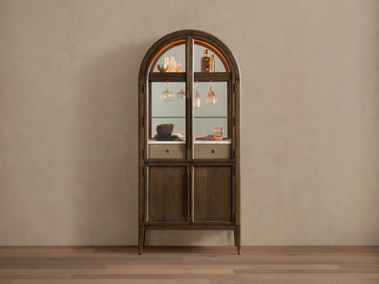 Hattie Bar Cabinet, dark wood with arched top. Lit glass doors reveal bottles, glasses, marble; solid lower doors.
