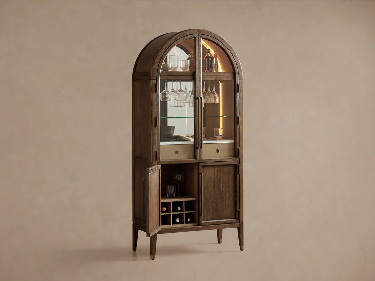 Hattie Bar Cabinet, dark wood with arched top. Lit glass doors reveal bottles, glasses, marble; solid lower doors.