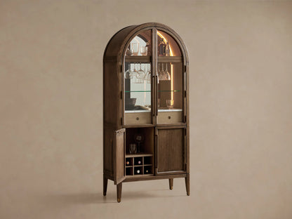 Hattie Bar Cabinet, dark wood with arched top. Lit glass doors reveal bottles, glasses, marble; solid lower doors.