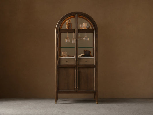 Hattie Bar Cabinet, dark wood with arched top. Lit glass doors reveal bottles, glasses, marble; solid lower doors.