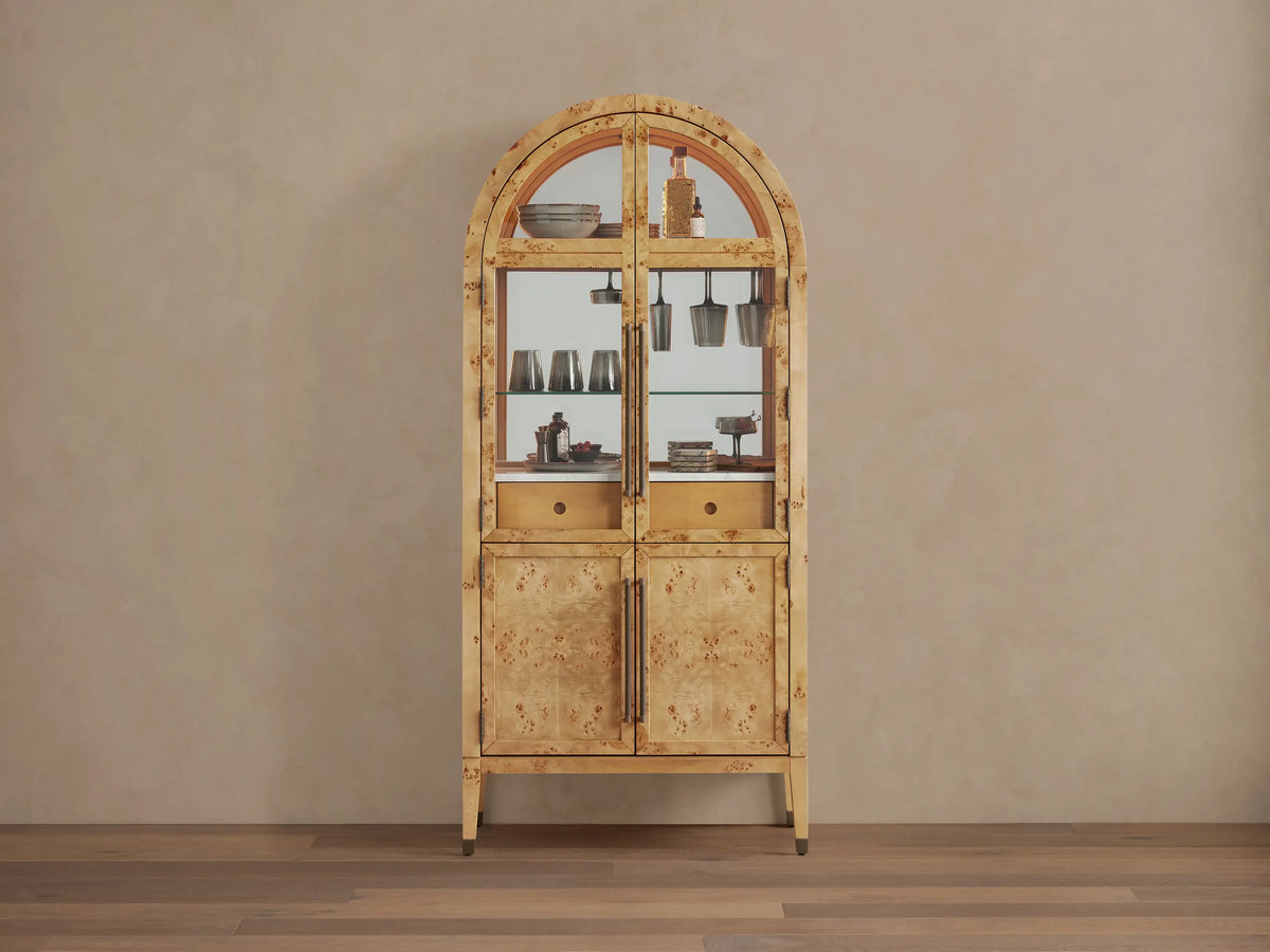Hattie Burl Bar Cabinet, light mappa burl wood with arched top, glass doors revealing barware, and solid lower doors with brass hardware.