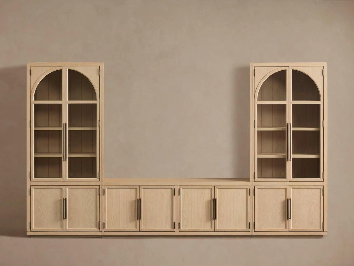 Hattie Glass Media Cabinet in light oak, angled. One bottom door open revealing interior shelf. Top glass doors show decor. Antiqued reeded hardware.