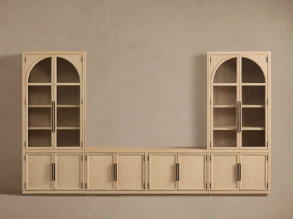 Hattie Glass Media Cabinet in light oak, angled. One bottom door open revealing interior shelf. Top glass doors show decor. Antiqued reeded hardware.