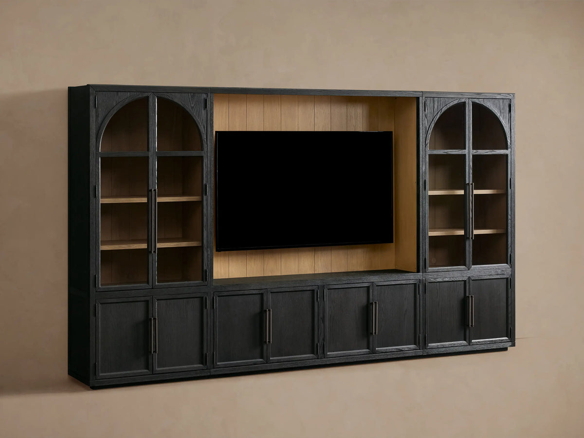 Front view of the dark oak Hattie Wall Unit, with a black TV screen, arched glass cabinets showing light wood shelves, and lower cabinets with reeded hardware.