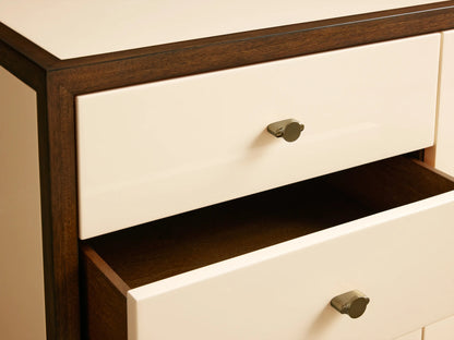 Henton Media Console with a wooden frame, three cream-colored drawers, and a sliding door. Brass handles and exposed shelves feature books and a pot.