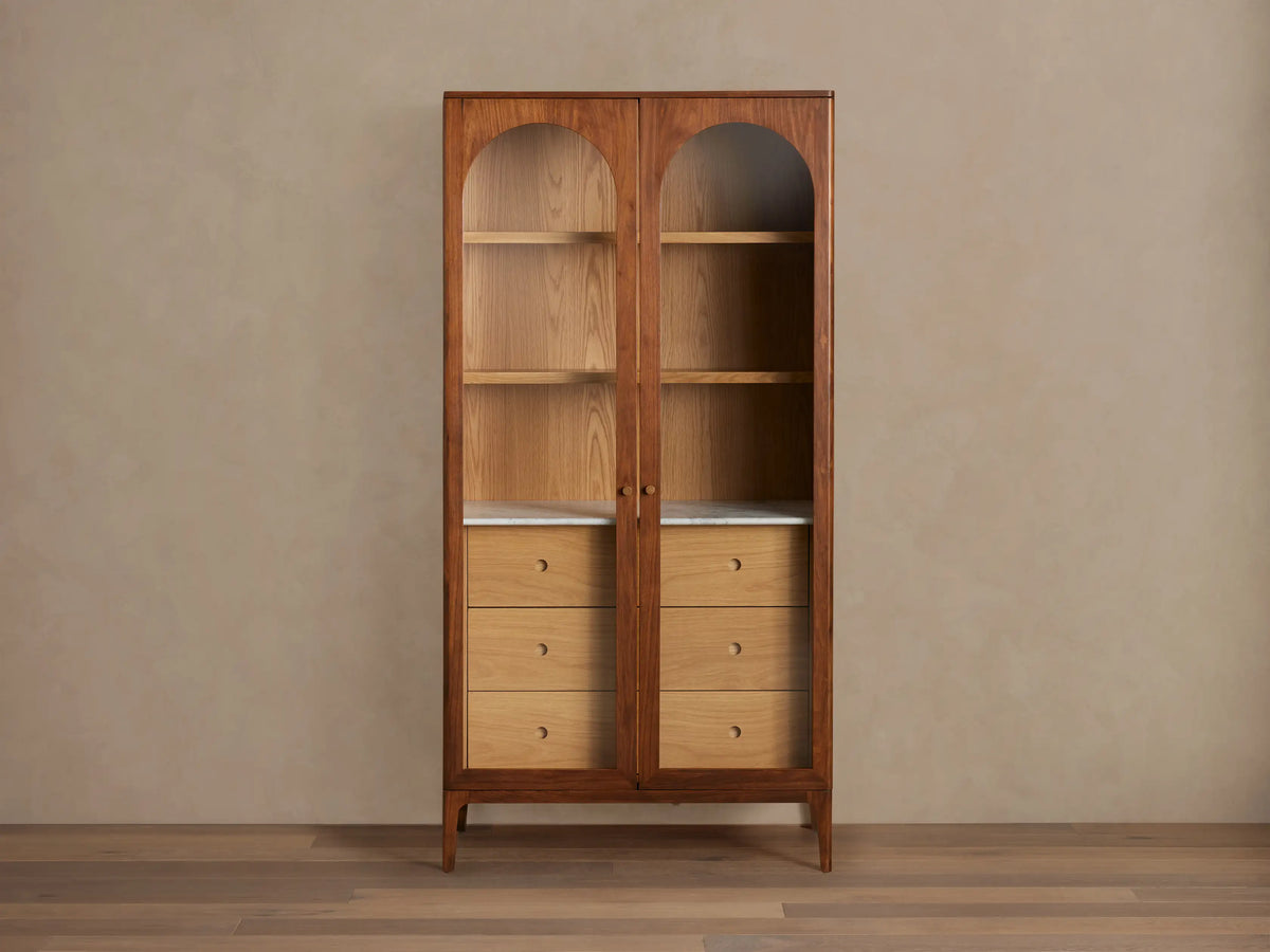 Close-up of the Linley Glass Cabinet showing the Carrara stone surface on top, with two beige books and a small bronze vase. The wood grain of the cabinet is visible with round cutout handles.