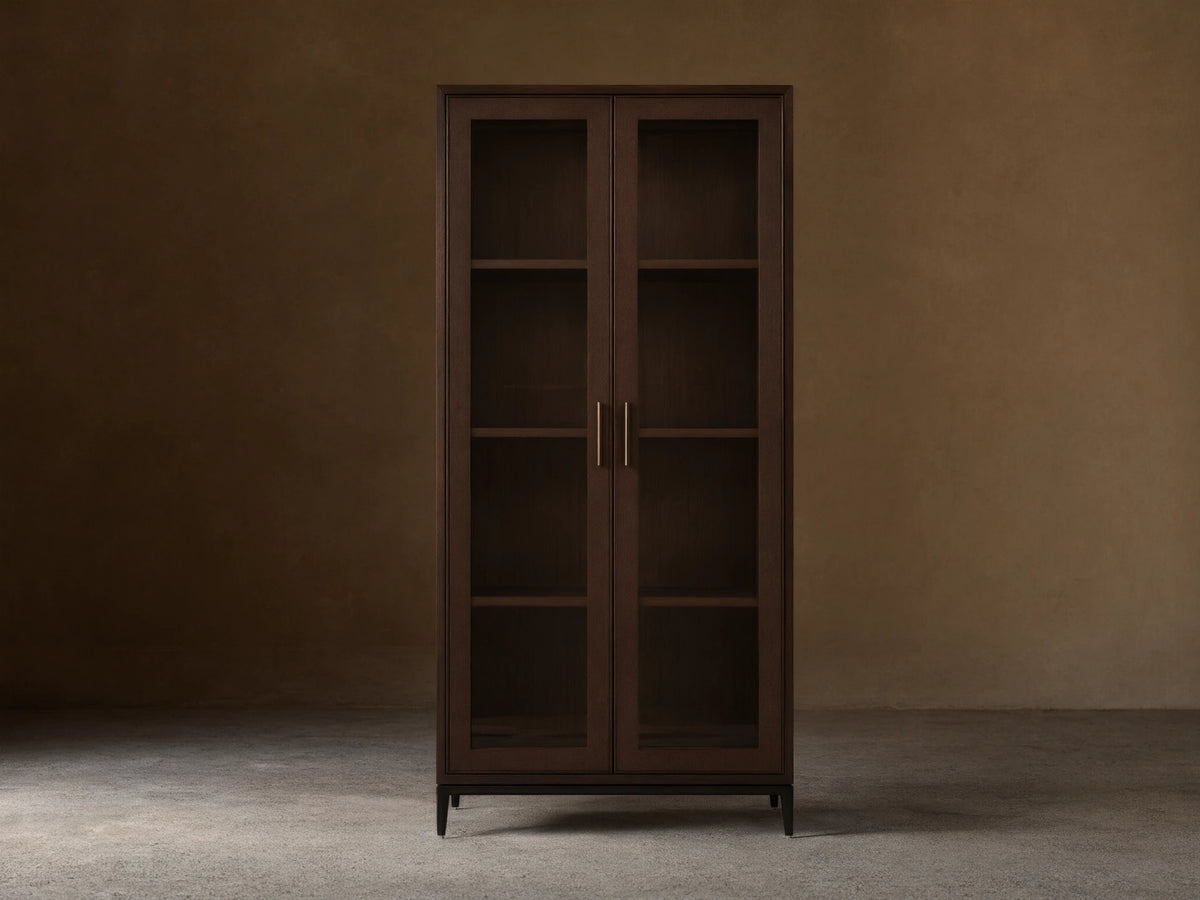 Malone Glass Cabinet in dark walnut finish with dual tempered glass doors, three shelves, and metallic handles, against a beige wall.