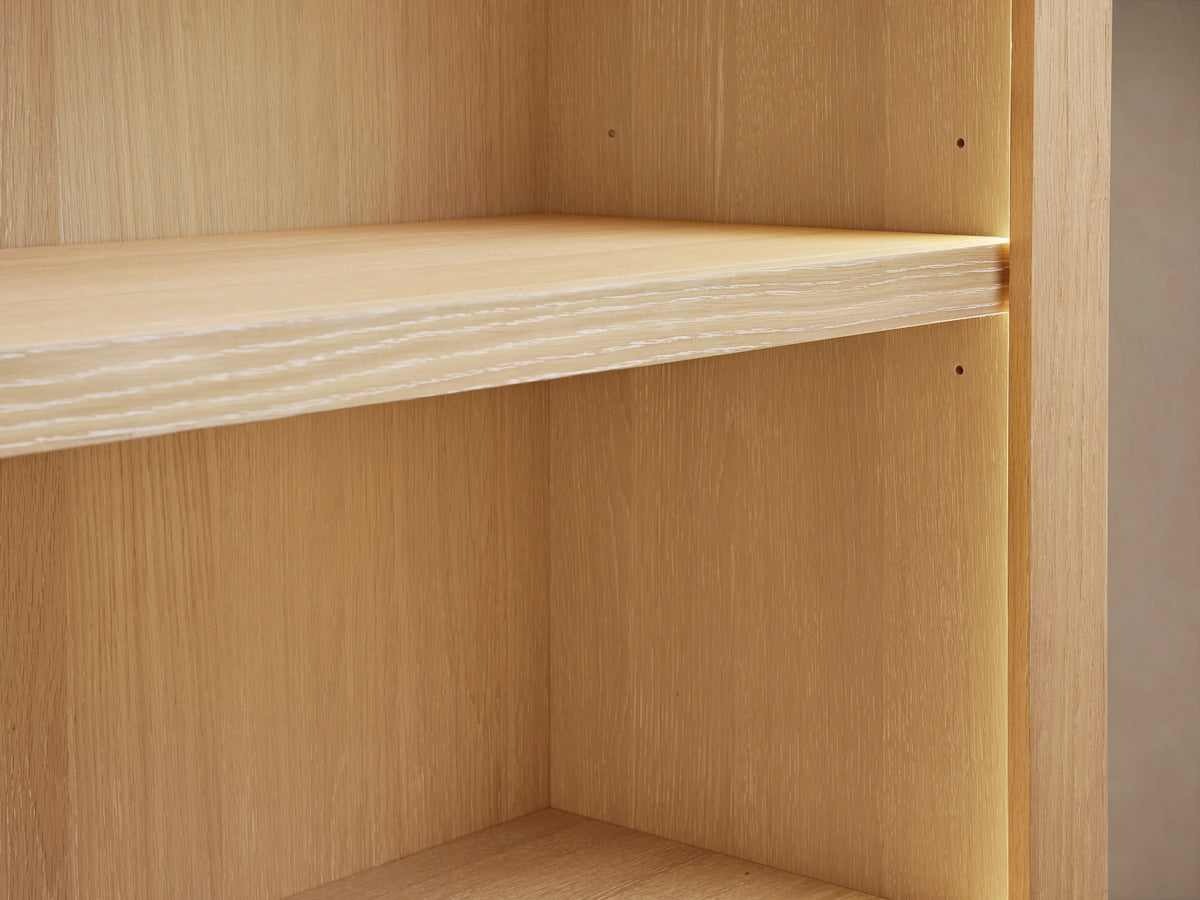 Nason Modular Bookcase crafted from light sand-finished white oak, featuring 6 empty shelves & a cord hole, standing against a beige wall.