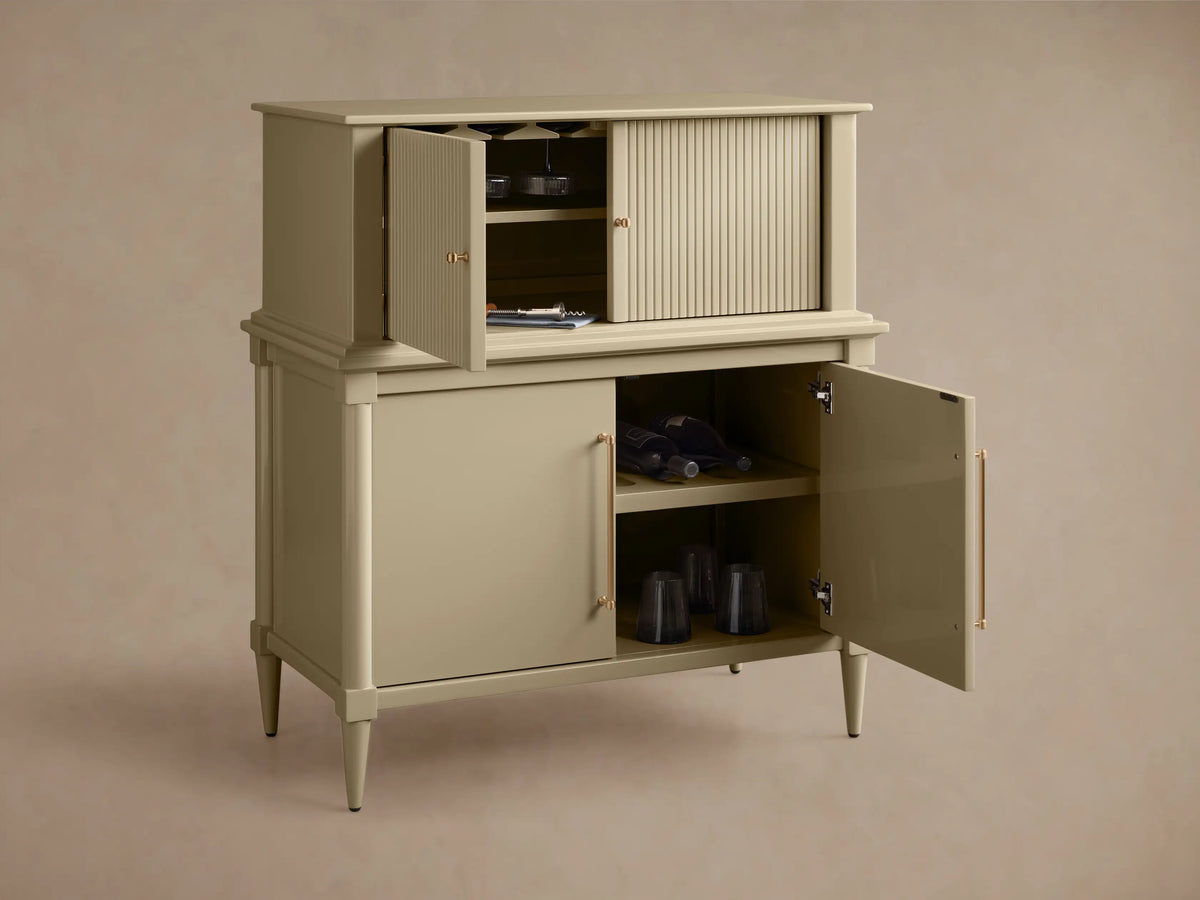 Angled front view of the light greige Oakleigh Bar Cabinet with reeded detailing. Upper doors ajar, lower doors with brass pulls open.