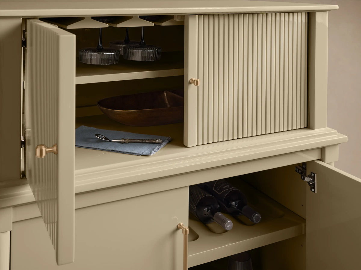 Interior view of the open light beige Oakleigh Bar Cabinet, revealing reeded doors, brass hardware, hanging stemware, and wine bottles.