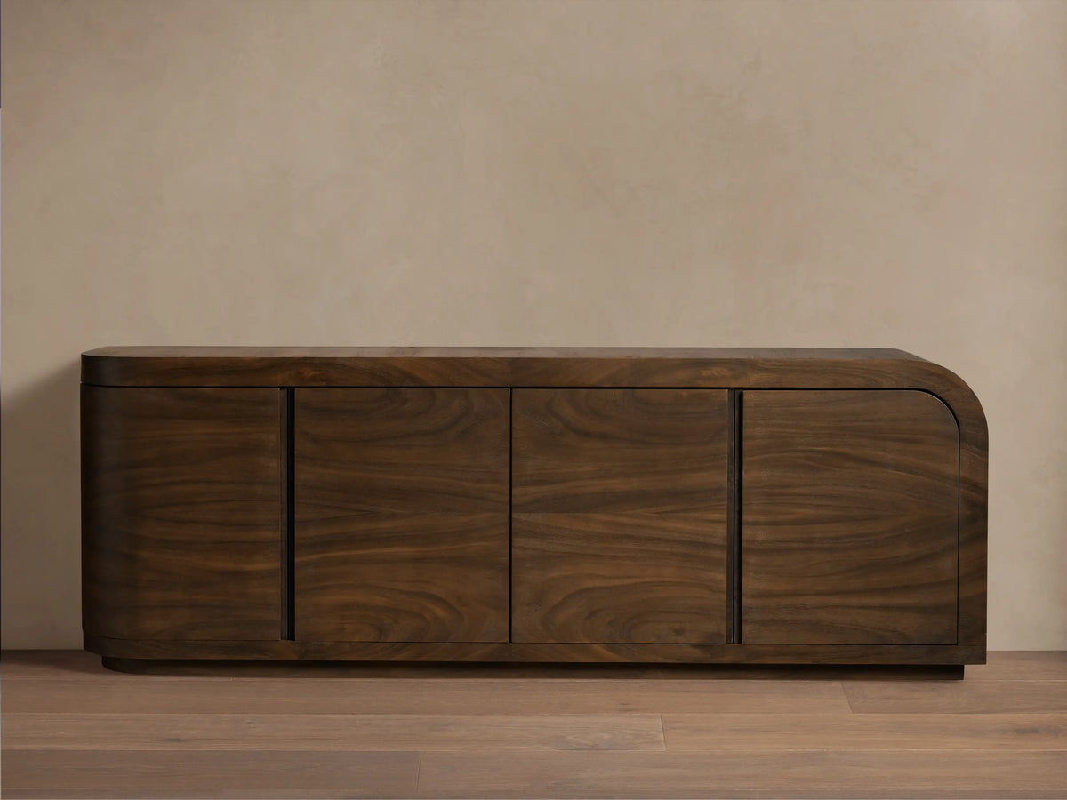 Dark brown guanacaste wood Pliego Sideboard with rounded right side, four doors, on wood floor against a light beige wall.
