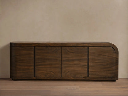 Dark brown guanacaste wood Pliego Sideboard with rounded right side, four doors, on wood floor against a light beige wall.