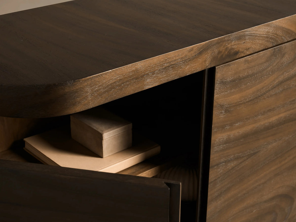 Close-up of Pliego Sideboard, dark guanacaste wood with visible grain. Its curved door is partly open, revealing a shelf, book, and box.