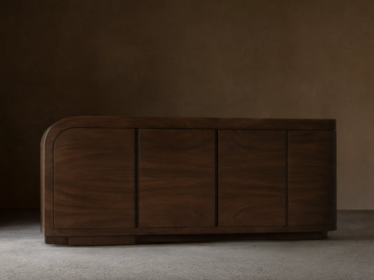 Dark brown guanacaste wood Pliego Sideboard with rounded right side, four doors, on wood floor against a light beige wall.