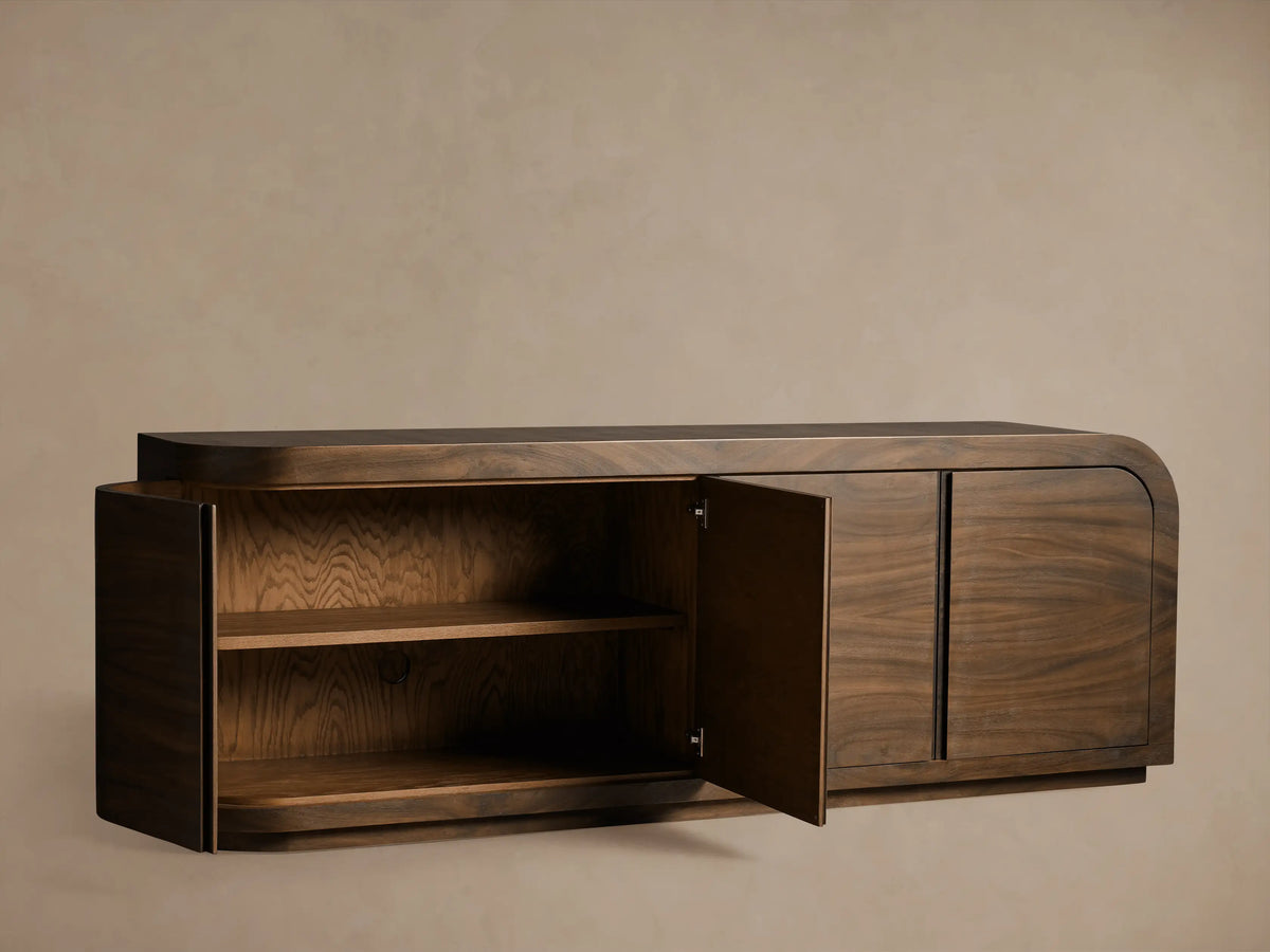 Dark brown Pliego Sideboard, left door open revealing two shelves. Curved design, rich guanacaste wood grain, floating against a beige wall.