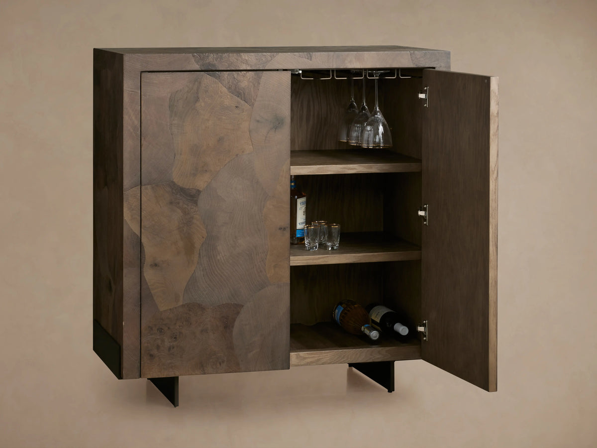 Polanco Bar Cabinet with one door open, revealing wine shelves, glasses, and bottles inside. Made of ash wood with natural graining and dark base.