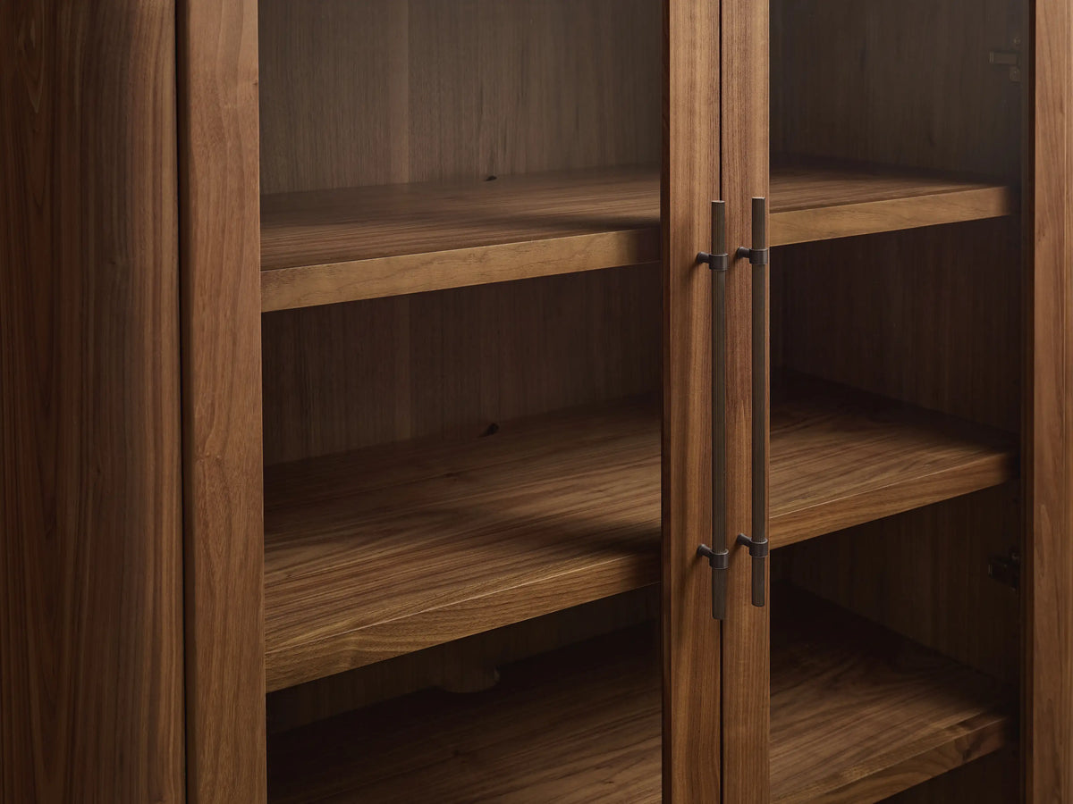 Close-up of Piermont Modular Glass Media Cabinet. Dark walnut wood, open lower door reveals shelves, bronze handles, plinth base.