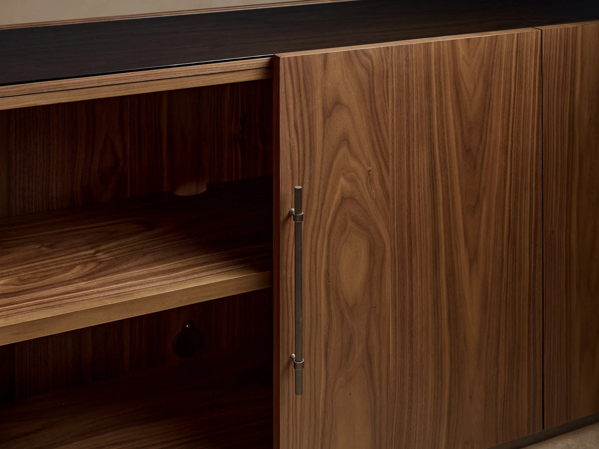 Close-up: Piermont Modular Glass Media Cabinet Set. Dark walnut wood with prominent grain, bronze handles, and plinth base. Glass sections visible.