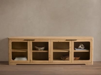Front view of a light natural wood cabinet with four glass doors, rounded corners, interior shelves, and dark metal handles.