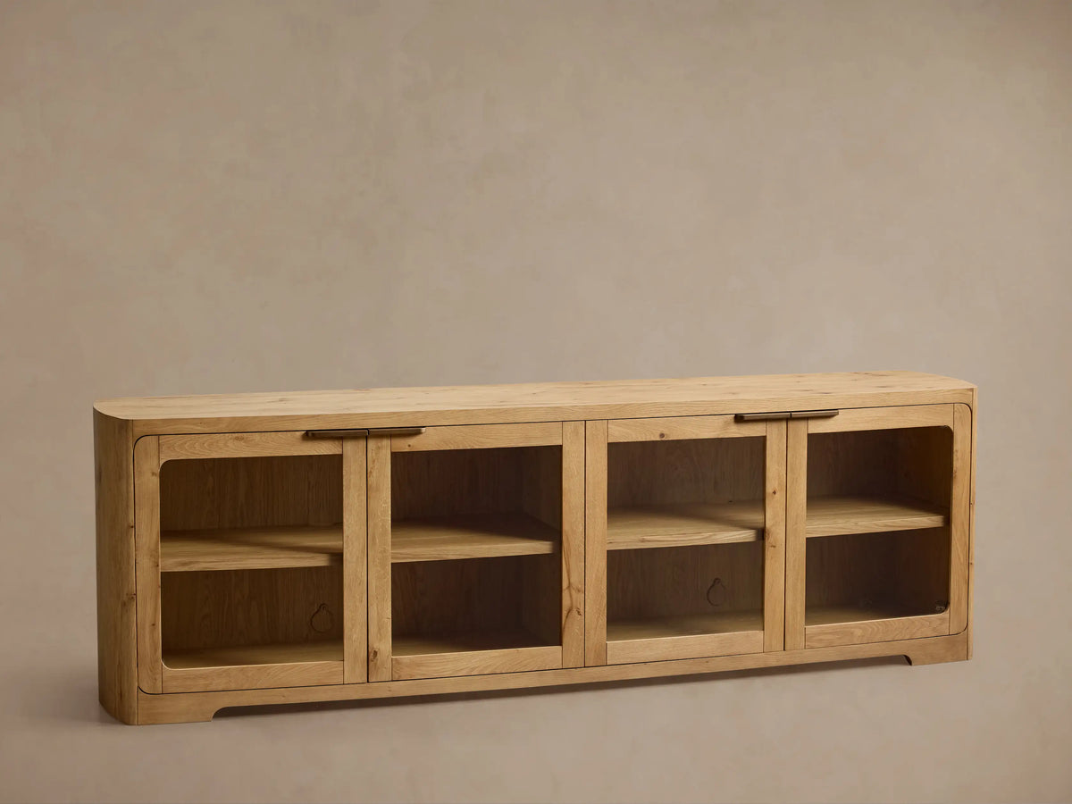 Front view of a light natural wood cabinet with four glass doors, rounded corners, interior shelves, and dark metal handles.