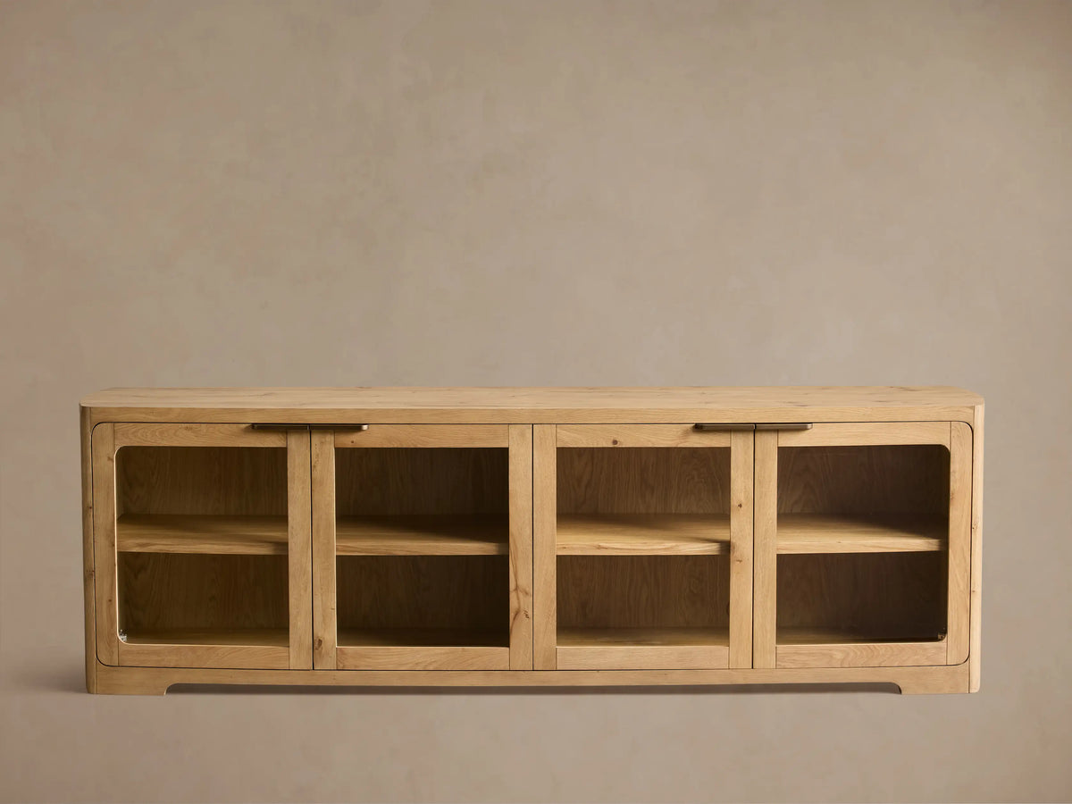 Close-up of light oak wood cabinet with visible grain, rounded top, glass doors, and brass bar handle.