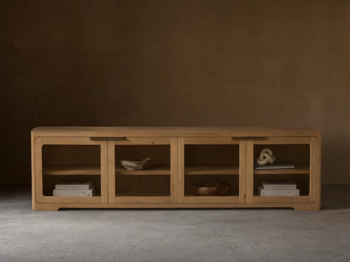 Front view of a light natural wood cabinet with four glass doors, rounded corners, interior shelves, and dark metal handles.