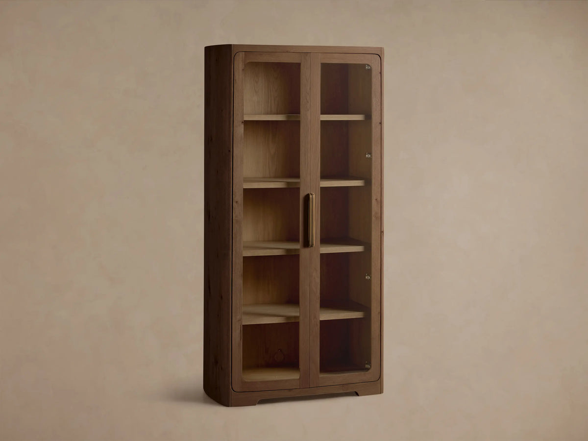Angled front view of the light brown Redding Glass Cabinet, featuring two glass doors, four shelves, and antique brass hardware.