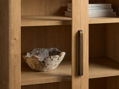 Close-up of the Redding Glass Cabinet's natural oak frame, glass door, and antique brass-finished handle, with a rustic bowl inside.